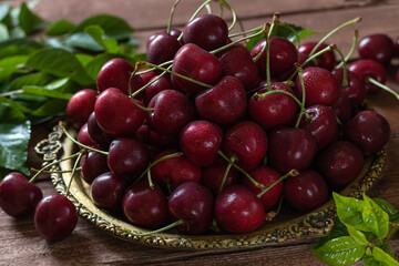 A metal vintage dish filled with cherries, cherry leaves nearby.