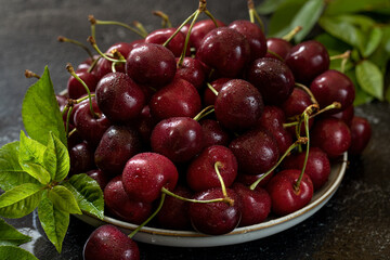 White ceramic dish filled with cherries, cherry leaves diagonally. Dark background.