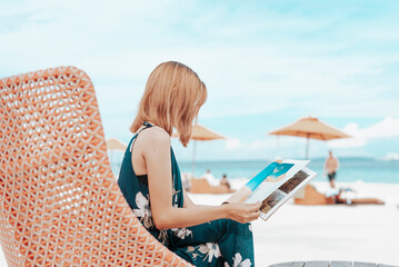 A young lady in a blue floral dress reading a lifestyle magazine while sitting on a comfy lounge...