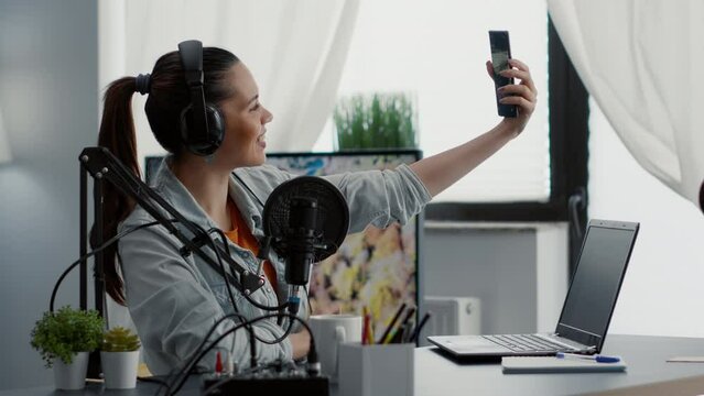 Famous internet influencer taking selfie photo with smartphone while streaming. Digital content creator sitting at home studio desk while taking pictures with touchscreen mobile phone.