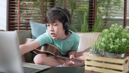 Little asian child boy online learning playing ukulele on armchair at living room with cheerful and fun singing along at home,online study social distance learning from home ideas concept - Powered by Adobe