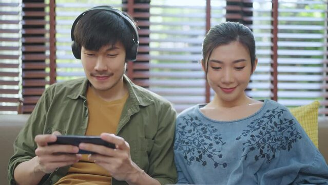lovely asian marry couple adult stay home lifestyle male playng game smartphone online with his wife smile laugh use laptop device sit relax together on sofa living room at home stay home concept