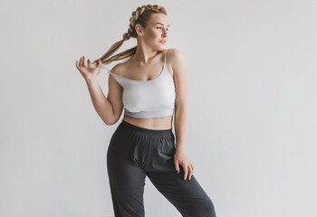 attractive young blond woman with curves posing after a workout in a sports top and yoga pants on a gray background. Plus size model goes in for sports and shows her body. Self acceptance concept
