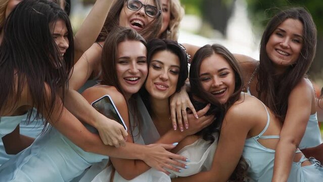 Pretty bridemaids in identical blue dresses run to hug bride together in park cheering waving hands. Woman party with friends on wedding day in elegant airy white gown and long veil. Slow motion