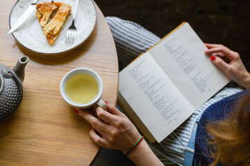 Woman's hands holding opened book, cup of tea pie on light wooden table.