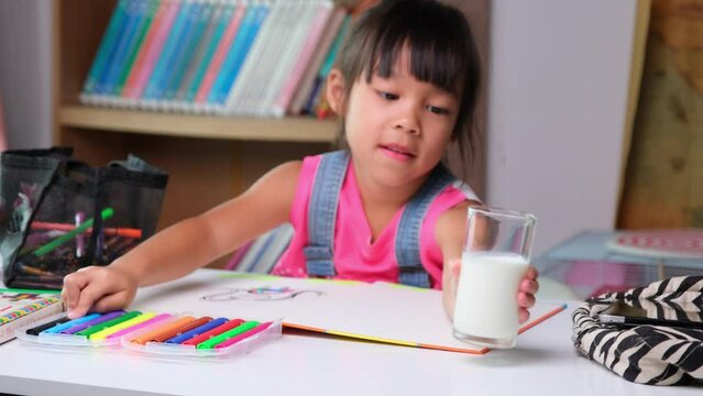 Cute little girl drinking milk while taking a break from drawing class. Asian little girl studying online and enjoying homeschooling. Child doing homework