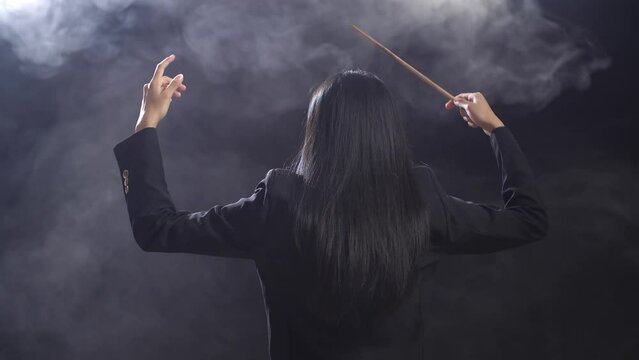 Back View Of Asian Conductor Woman Holding A Baton And Showing Gesture In The Black Studio With Fog 
