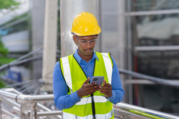 black engineer with glasses wear glow shirt and helmet, using mobile phone while working at construction site outside building in city