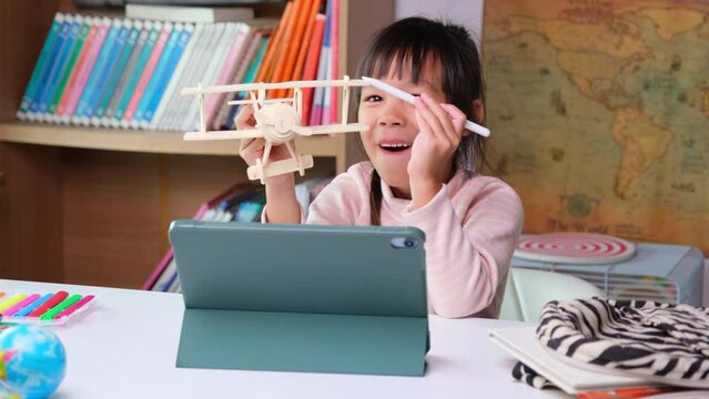 Cute little girl holding a Stylus pen working on a tablet and holding wooden model airplane learning from home with school teacher teaching online. Modern kid and education technology.