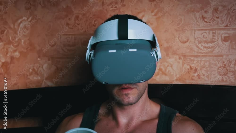 Poster man in a virtual reality helmet controls the controllers. a young man at home sitting on a bed is in
