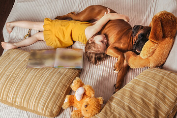a little girl in a yellow dress at home on the couch goes to bed with her big dog German boxer, next to a book and soft toys
