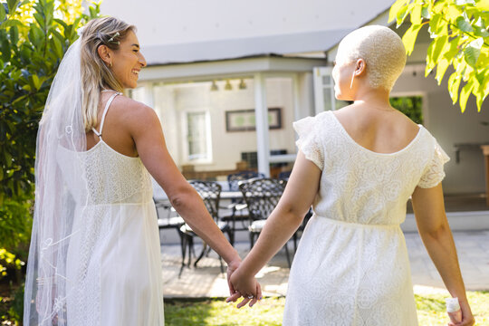 Image of happy diverse lesbian couple in wedding dresses holding hands
