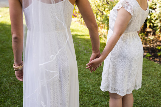 Image of midsection diverse lesbian couple in wedding dresses holding hands