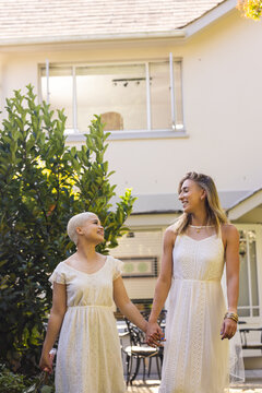 Vertical image of happy diverse lesbian couple in wedding dresses holding hands