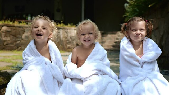 Three kids laugh wrapped in a white towel after taking a dip in the pool while on vacation. The contagious laughter of children who are washed away in the frame. Vacation concept with children.