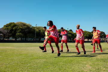 Fototapeta na wymiar Male multiracial players wearing red uniforms running and exercising against clear sky in playground
