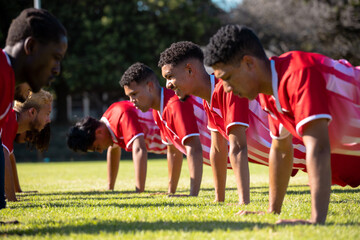 Male multiracial players in red uniforms practicing push-ups in a row on grassy field at playground