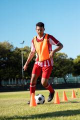 Biracial male player in red jersey running with soccer ball between cones against clear sky