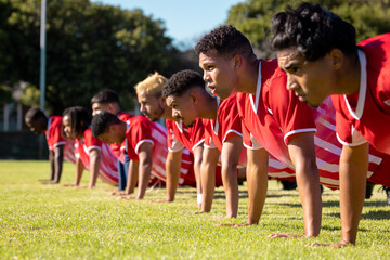 Male multiracial players wearing red uniforms in a row doing push-ups on grassy playground