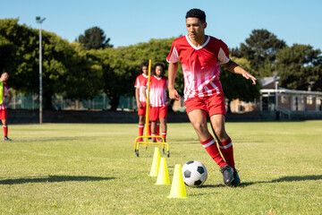 Multiracial male players in red jersey running with soccer ball between cones at playground