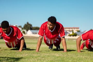 Multiracial male players in red uniforms doing push-ups on grassy playground against clear sky