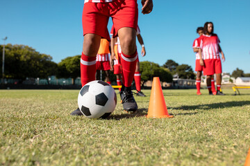 Low section of male multiracial players dribbling ball between cones at playground against clear sky