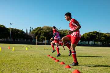 Multiracial male players in red jersey jumping by disc cones on grassy playground against clear sky