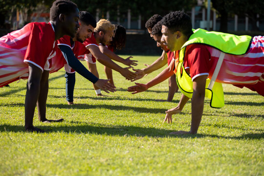 Multiracial male players in red uniforms practicing high five push-ups in a row on grassy field