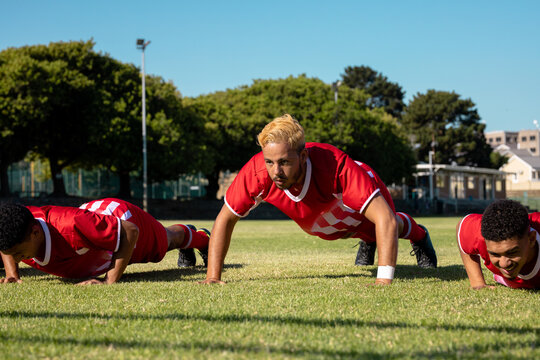 Male multiracial players in red uniforms practicing push-ups on grassy field against clear sky