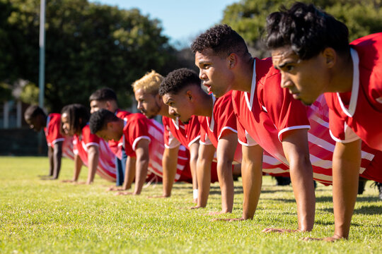 Male multiracial players wearing red uniforms in a row doing push-ups on grassy playground