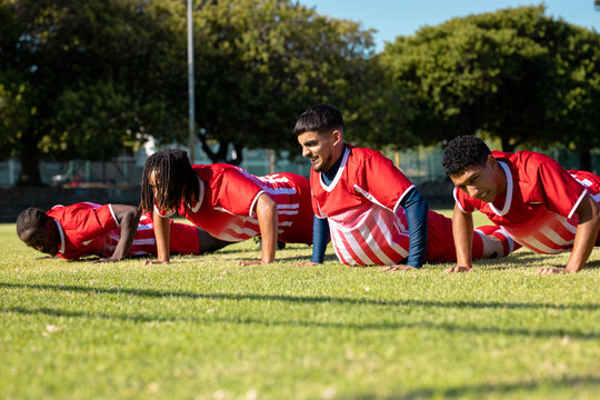 Male multiracial players wearing red uniforms doing push-ups on grassy playground in summer