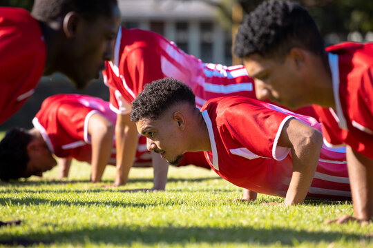 Multiracial male players wearing red uniforms practicing push-ups on grassy field in playground