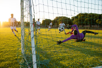 Multiracial goalkeeper defending ball of opponent goal during match against clear sky at sunset