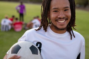 Close-up portrait of biracial male player with dreadlocks holding soccer ball in playground