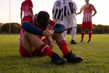 Caucasian sad male athlete sitting on grassy land with team players in background at playground - obrazy, fototapety, plakaty