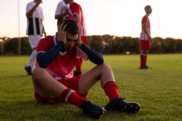 Caucasian sad athlete with head in hands sitting on land with players in background at playground