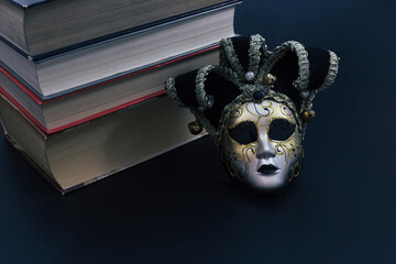 A stack of old books and a Venetian theatrical mask on a black background. Gloomy atmosphere