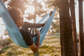 Handsome young man in glasses lying in hammock and relaxing with coffee. Forest travel.