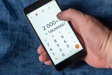 A man calculates his monthly loan payment on his smartphone. The loan amount is divided into twelve parts. The hand with the device is across the blue jeans.