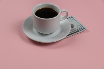 Espresso coffee and money against a pink background. A hundred-dollar bill lies under a white cup and saucer. A tip for the waiter. Close-up. Selective focus.