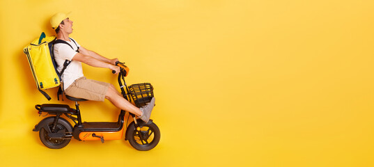 Full length side view portrait of man courier with thermo backpack in white T-shirt and cap riding electric bike, braking, avoids accident, looks scared and shocked, isolated over yellow background.