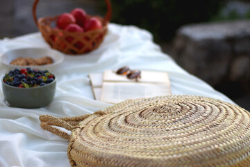 Fototapeta na wymiar Picninc blanket with straw bag, bowl of strawberries and blueberries, bowl of chocolate chip cookies, books, sunglasses and basket of apples in the garden. Selective focus.