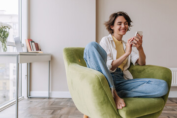 Caucasian brunette woman smiling at phone . Full view of pretty young woman holding and chatting on smartphone in home. Technology, lifestyle 