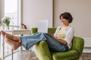 Beautiful caucasian woman using laptop at home . Full view of short hair woman sitting on chair...