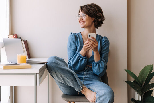Picture of attractive woman smiling to window . Beautiful young brunette woman sitting on chair at home with phone look away. Concept of working home, technology 