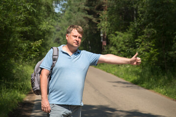 Fototapeta premium An adult tired blond man with a heavy backpack stands on the road and catches the car with a hand gesture.