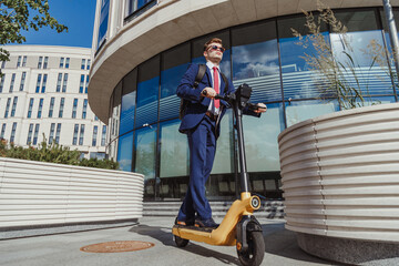 Young man in suit rides on electric scooter in business district at modern corporate building.