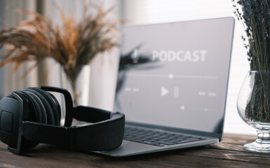There is a laptop on a wooden table with an audio podcast running on the screen. Headphones are lying nearby. Selective focus on audio headphones.