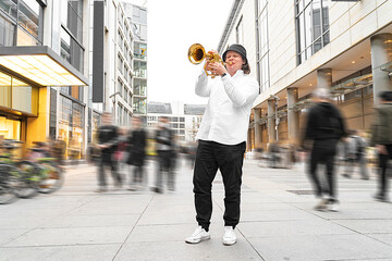 Young caucasian long-haired blond man in white shirt and hat playing funky jazz on golden trumpet with pleasure standing in middle of crowded modern downtown street with department stores, in Germany
