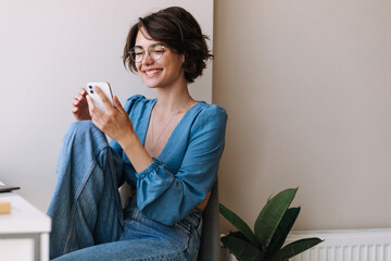 Charming woman holding phone at home . Caucasian smiling brunette woman looking and chatting on...
